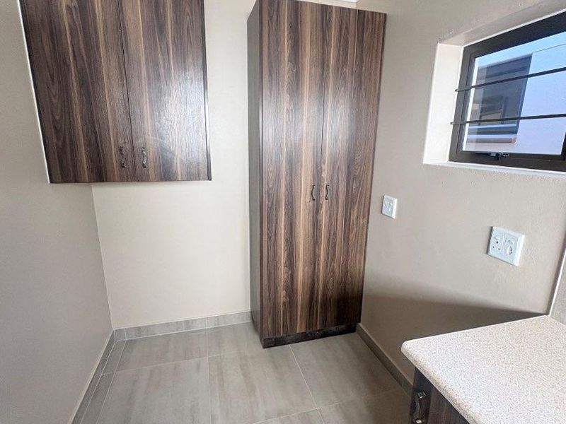 A clean, empty laundry area featuring modern wood-finish built-in cabinetry, tiled flooring, and a window with security bars.