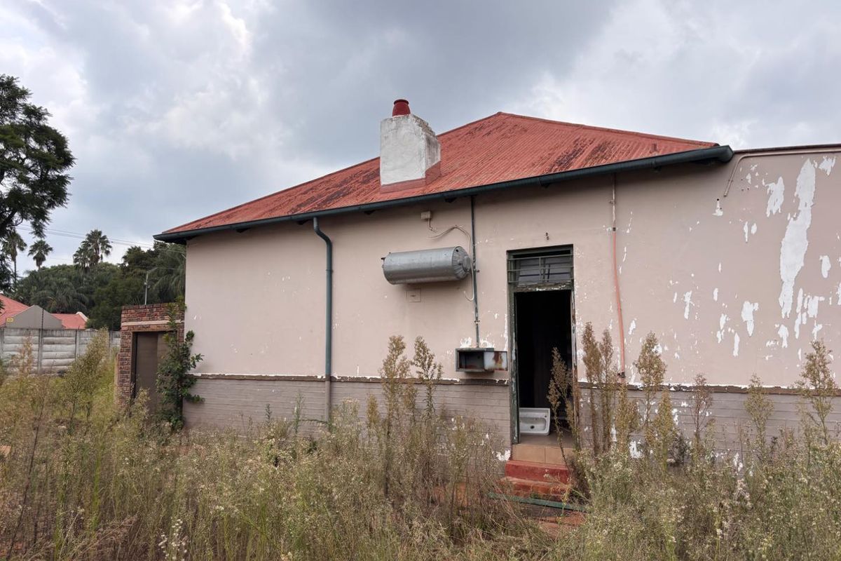 Rear exterior view of a house showing significant wear, including peeling paint, a rusted roof, and overgrown garden area requiring maintenance.