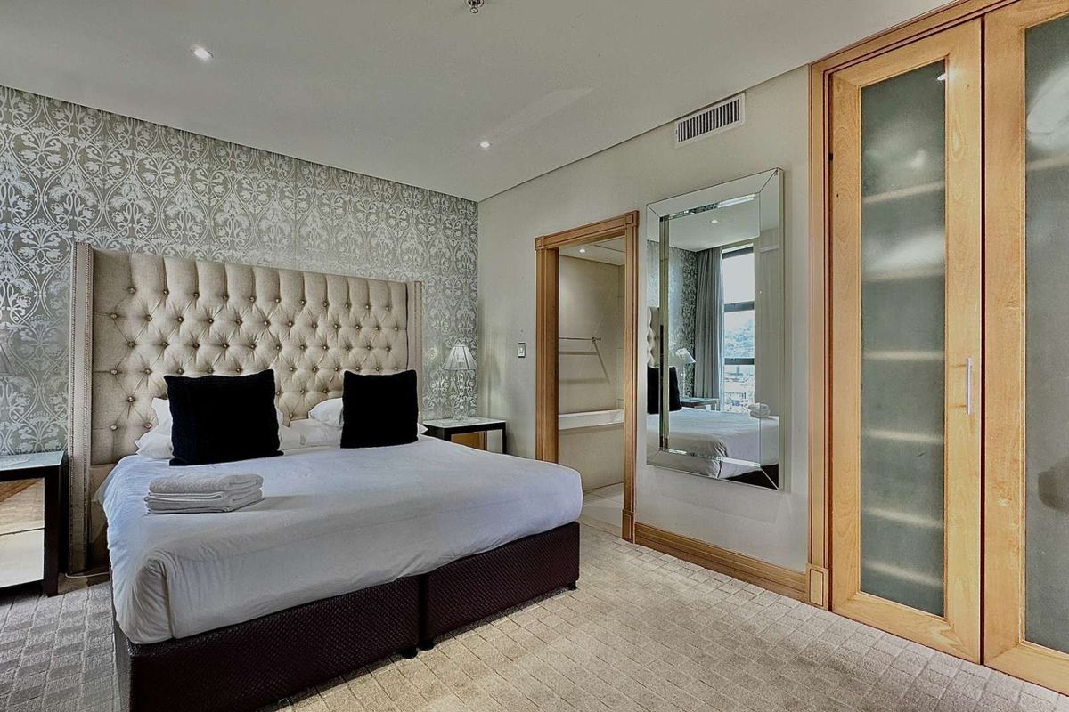 A well-appointed bedroom featuring a tufted headboard, patterned feature wall, integrated air conditioning vent, and built-in wooden wardrobe with frosted glass panels.