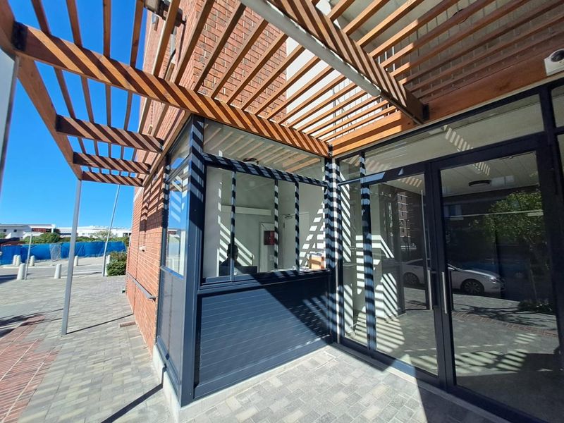 Modern building entrance featuring a timber pergola, brick facade, and glass storefront with paved exterior access.