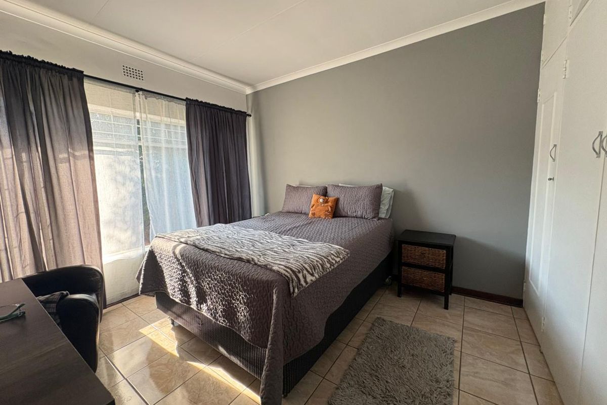 A bedroom featuring tiled flooring, built-in cupboards, and large windows with curtains, providing natural light and a neutral color palette.