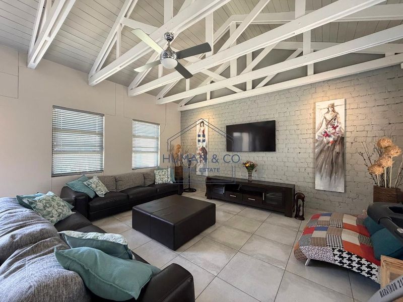 Spacious living room featuring a high vaulted ceiling with exposed white timber trusses, tiled flooring, and a characterful exposed brick feature wall.