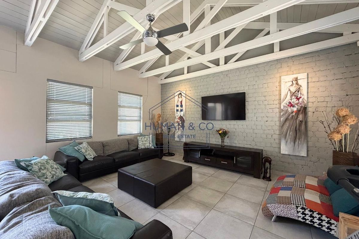 Spacious living room featuring a high vaulted ceiling with exposed white timber trusses, tiled flooring, and a characterful exposed brick feature wall.