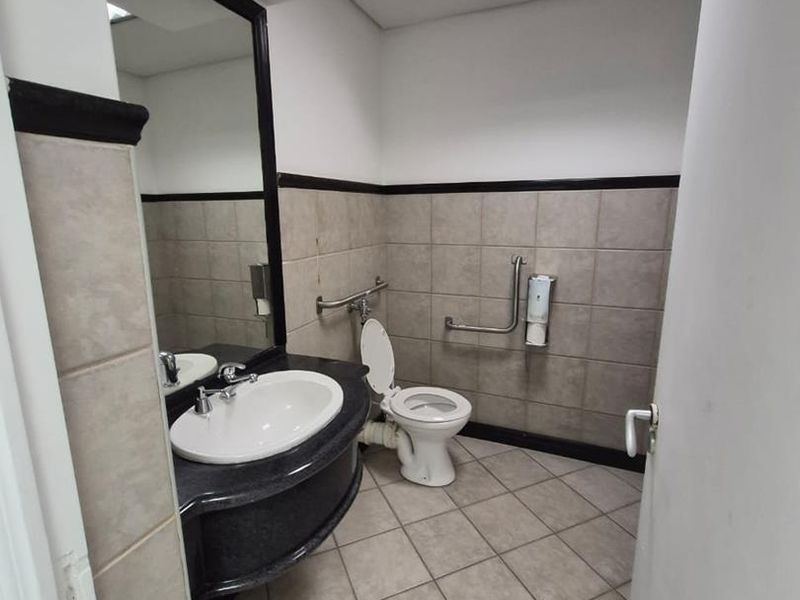 A functional office bathroom featuring a vanity basin, wall-mounted grab rails, and neutral ceramic tiling throughout.