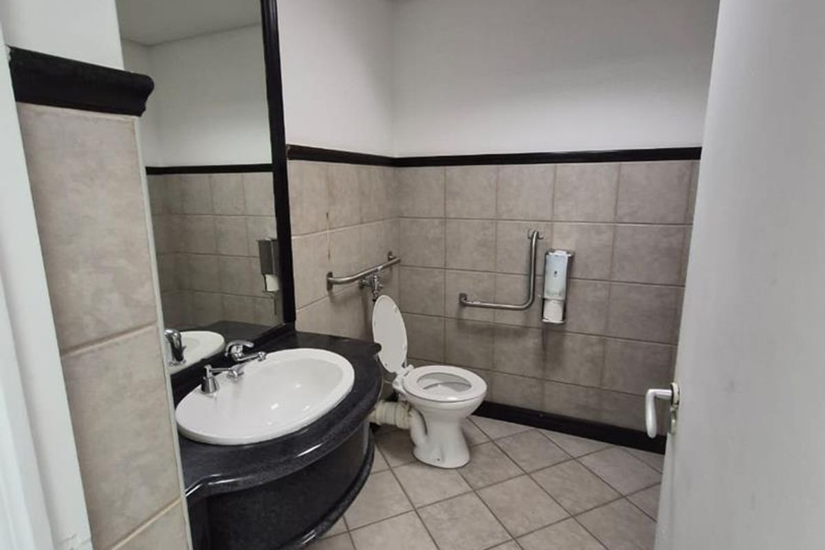 A functional office bathroom featuring a vanity basin, wall-mounted grab rails, and neutral ceramic tiling throughout.