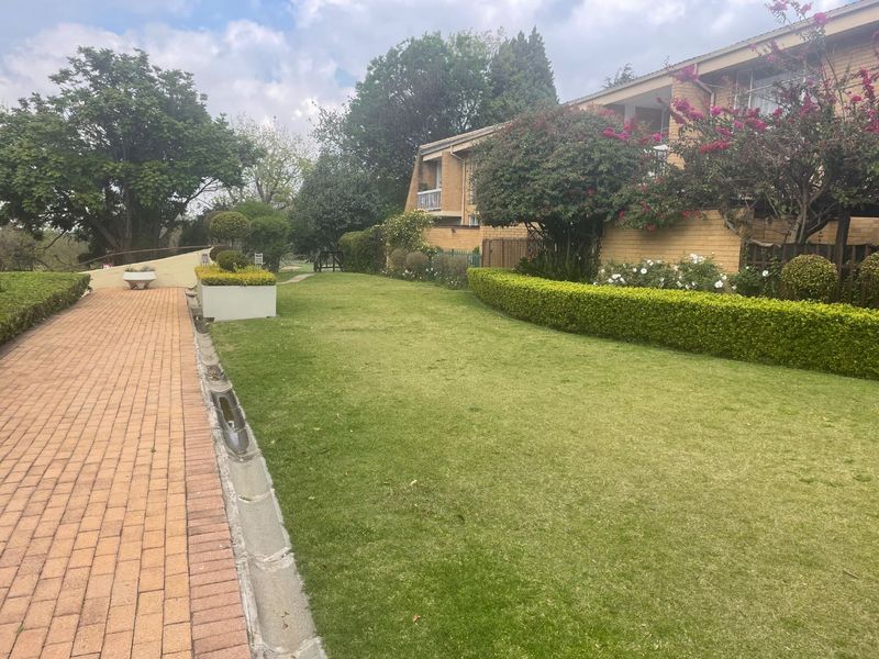 A well-maintained communal garden area featuring a brick paved walkway, manicured lawn, and established hedges alongside the apartment building exterior.