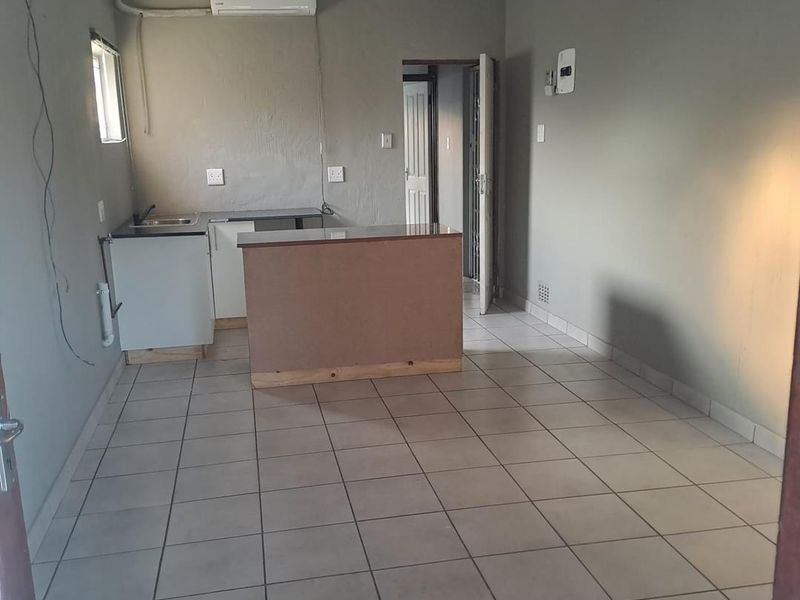 An open-plan living and kitchen area featuring tiled flooring, a small kitchenette with a sink, and a wall-mounted air conditioning unit.