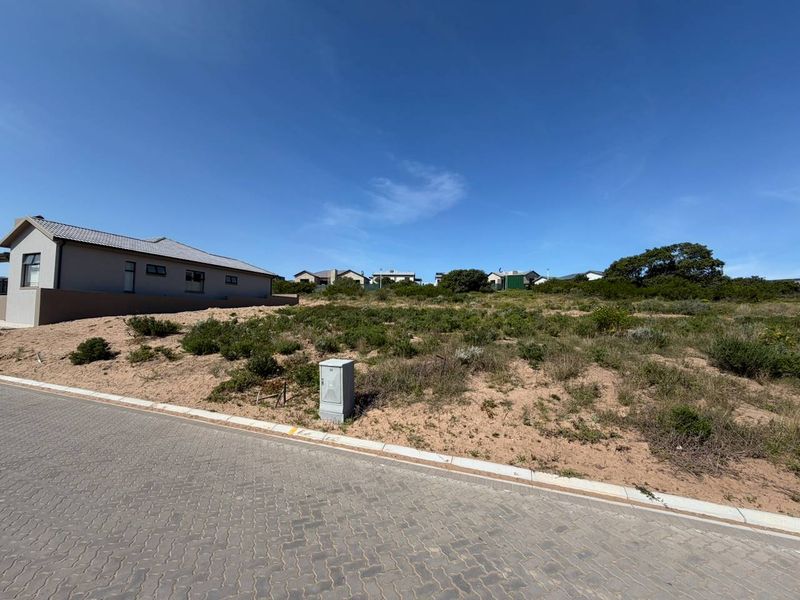 A vacant residential plot featuring natural vegetation, paved street access, and an existing electrical utility connection point.
