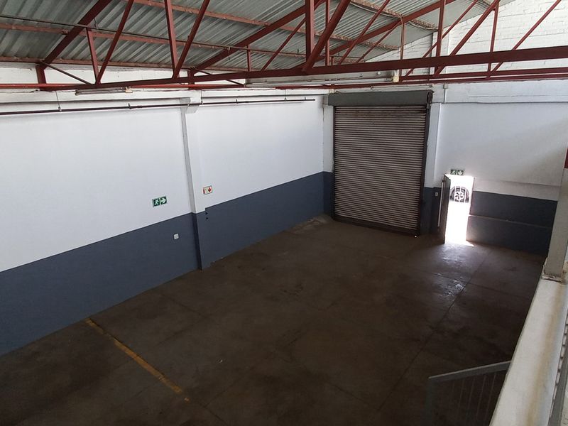 Spacious industrial warehouse unit featuring high ceilings with exposed steel trusses, a large roller shutter door, and a mezzanine level access point.