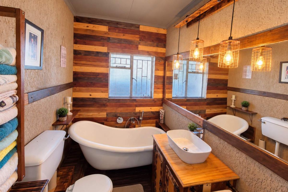 A rustic-style bathroom featuring a freestanding bathtub, vessel sink on a timber vanity, large mirror, and warm pendant lighting against textured and wood-clad walls.