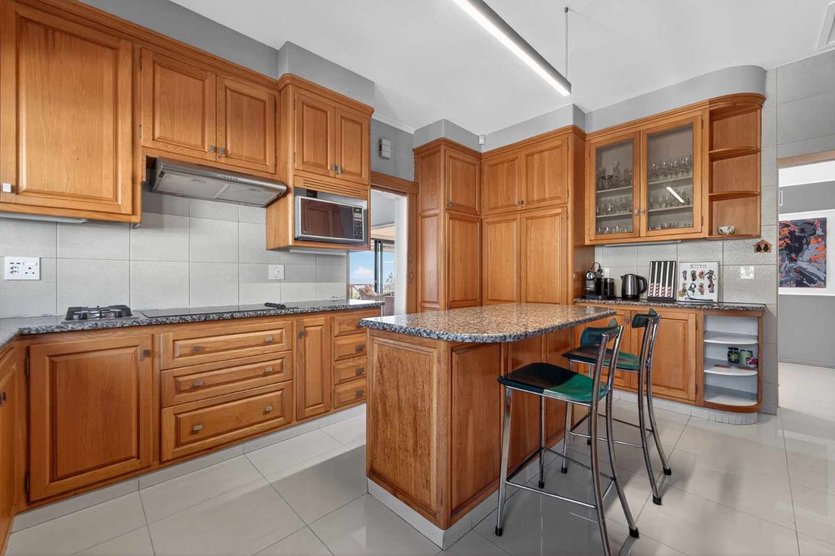 A functional kitchen featuring extensive wooden cabinetry, granite countertops, a central island, and integrated appliances, finished with neutral wall tiles and floor tiles.