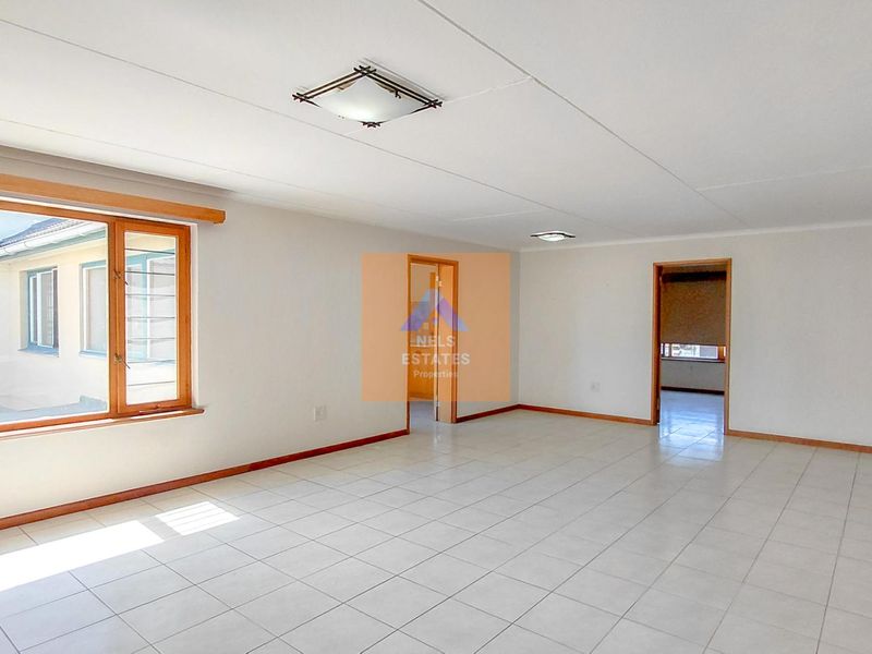 A spacious, empty living area featuring neutral tiled flooring, wooden window frames, and multiple doorways leading to adjacent rooms.