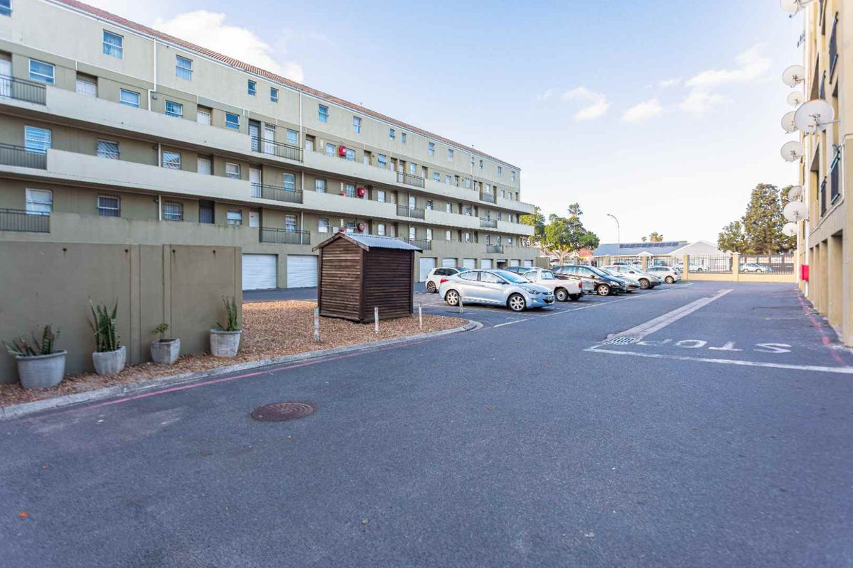 Exterior view of an apartment complex featuring a paved parking area, multi-story residential building with balconies, and designated parking bays.