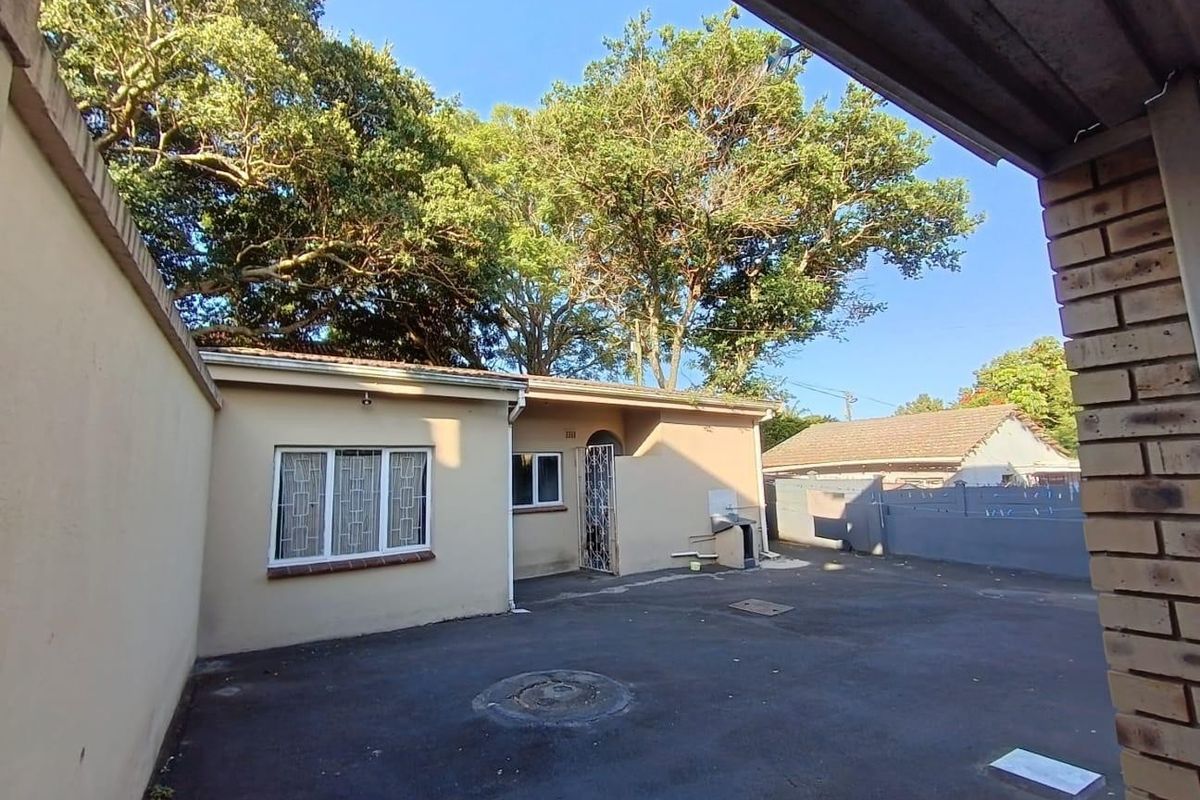A view of the rear exterior area featuring an asphalt driveway, brick boundary walls, and mature trees providing shade to the property.