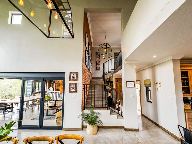 Spacious open-plan living area featuring double-volume ceilings, exposed brick accents, a modern staircase, and seamless flow to the outdoor patio through large sliding doors.
