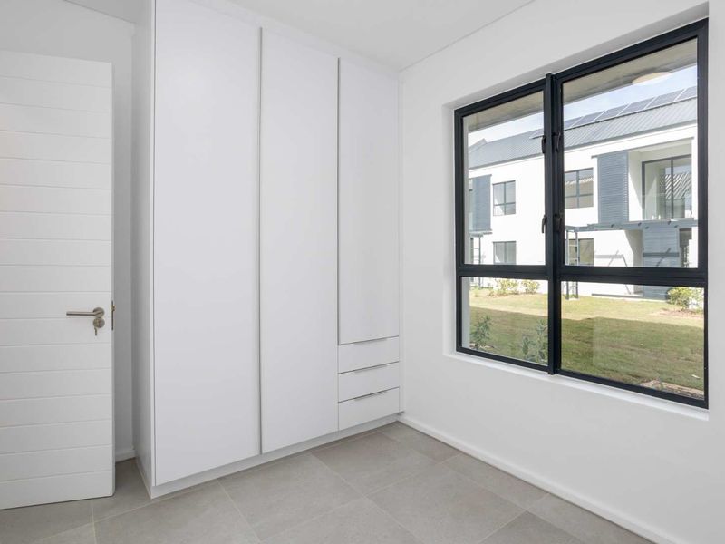 A bright, modern bedroom featuring neutral tiled flooring, a large built-in wardrobe with clean lines, and a black-framed window overlooking the complex grounds.