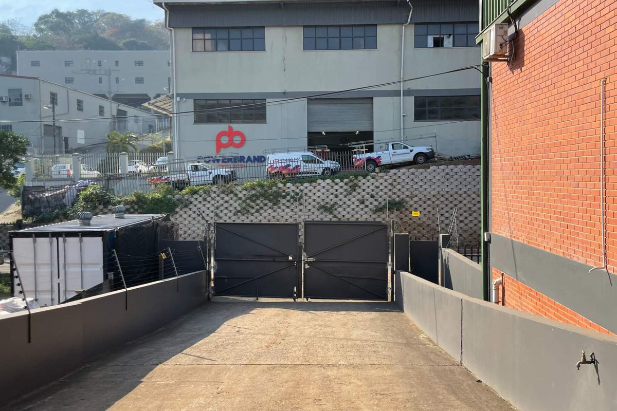 Rear exterior view of the industrial property featuring a concrete driveway, secure metal gate, and retaining wall structure.