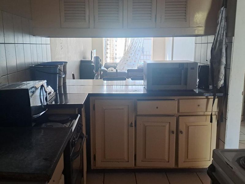 A functional kitchen featuring dated built-in cabinetry, tiled backsplash, and tiled flooring, with a pass-through window overlooking the living area.