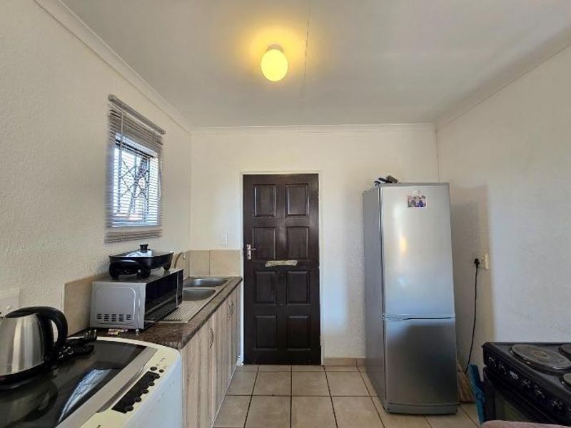 A compact kitchen featuring built-in cabinetry, laminate countertops, a stainless steel sink, and tiled flooring, with a window fitted with security bars.