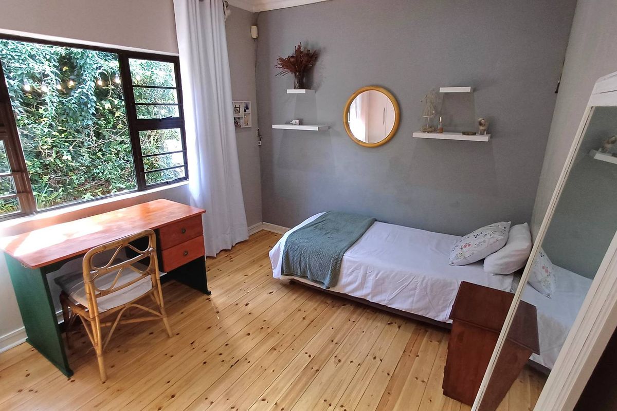 A well-lit bedroom featuring polished timber flooring, a large window with garden views, and neutral grey wall finishes.