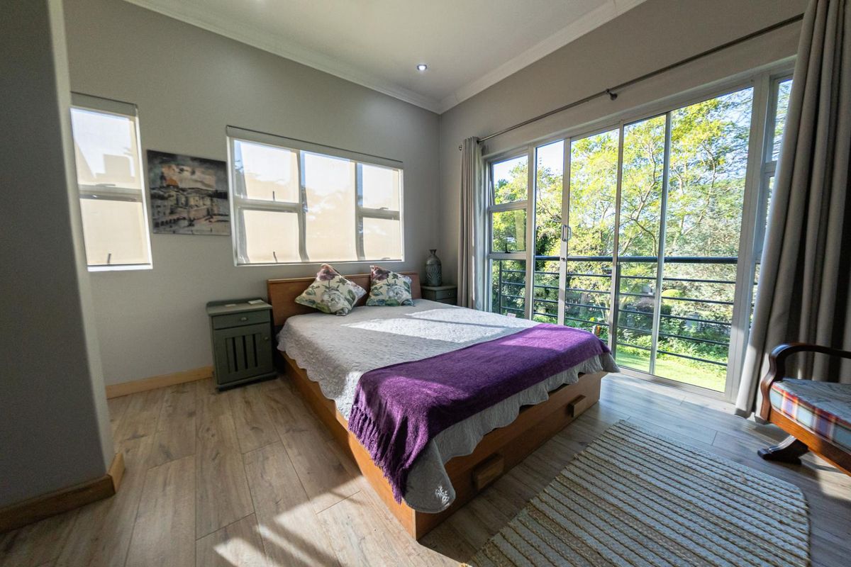 A bright bedroom featuring wood-look flooring, large windows, and sliding glass doors that open onto a balcony with lush garden views.