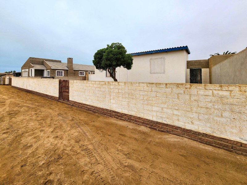 Exterior view of the property featuring a long boundary wall with a timber gate, set against an unpaved sandy approach.