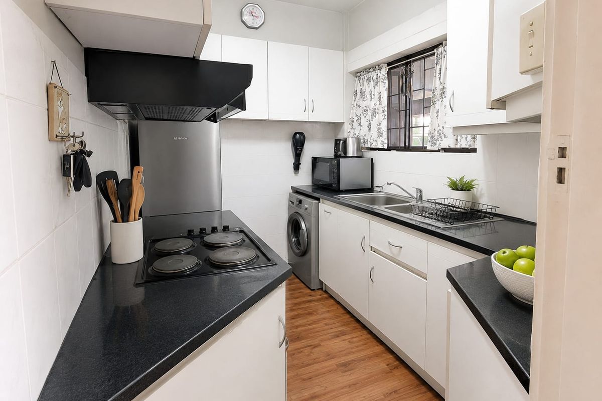 A functional galley-style kitchen featuring white cabinetry, dark laminate countertops, an electric hob with extractor fan, and integrated laundry plumbing.