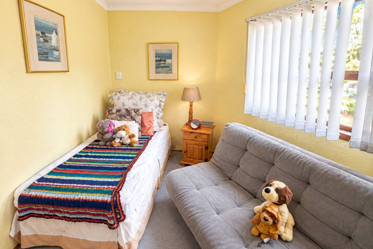A compact bedroom featuring neutral yellow walls, carpeted flooring, and vertical window blinds, offering a functional space for a single bed and seating.