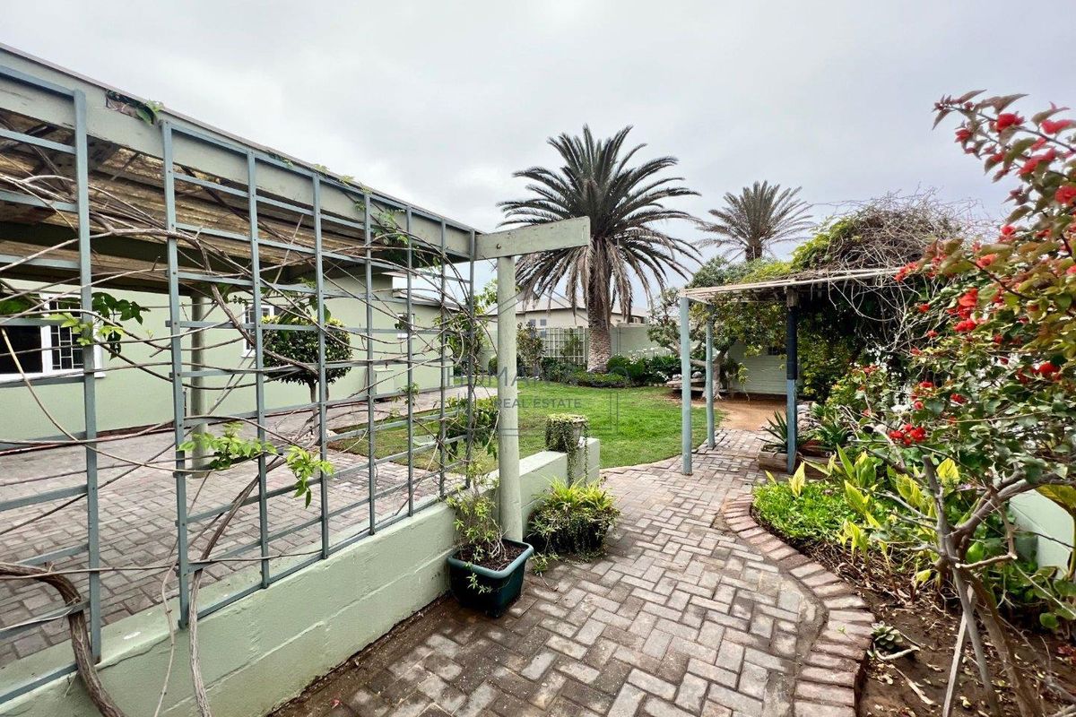 A landscaped rear garden featuring paved walkways, a metal pergola structure, and established garden beds with mature trees.