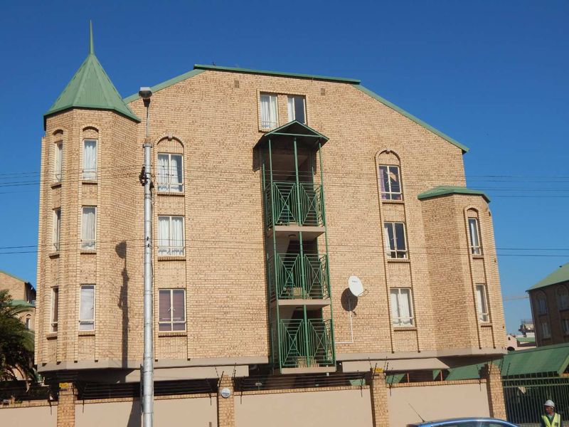 Front elevation of a multi-story apartment building featuring face brick construction, green metal roofing, and integrated balcony structures with security fencing.