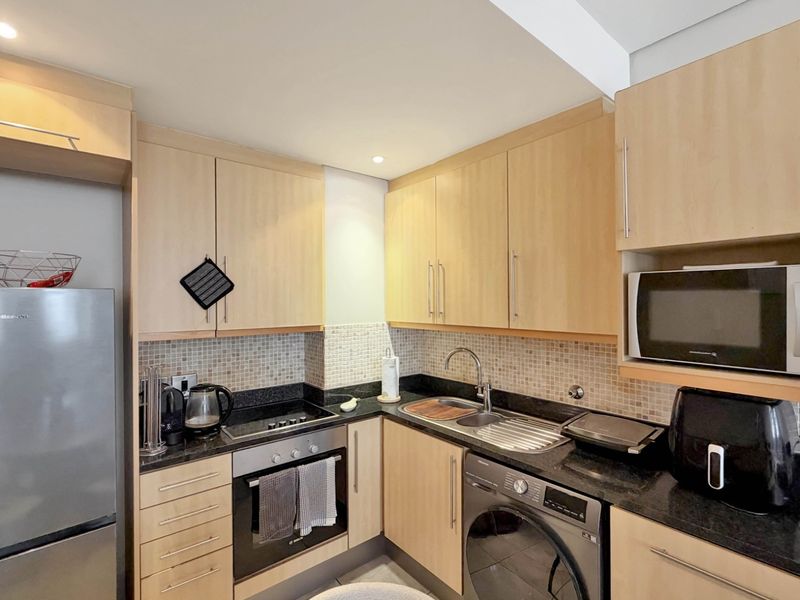 Compact kitchen featuring light wood cabinetry, granite countertops, mosaic tile backsplash, and integrated appliances including an oven, hob, and washing machine.