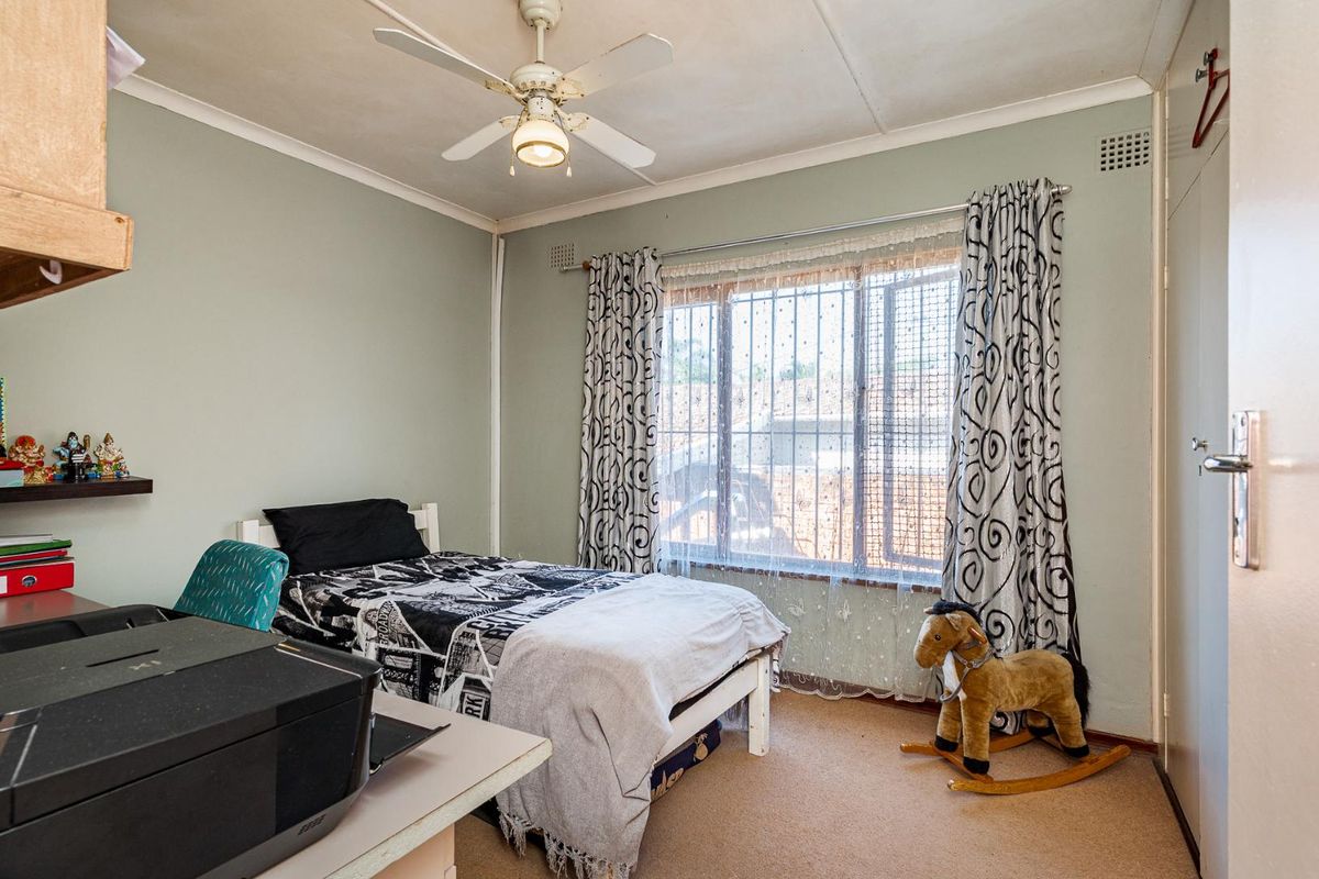 A functional bedroom featuring built-in cupboards, a ceiling fan, and security bars on the window, providing a comfortable and secure living space.