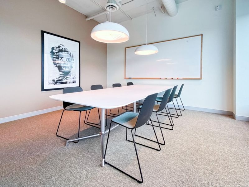 A modern office meeting room featuring a large whiteboard, pendant lighting, exposed ceiling ducting, and neutral carpeted flooring.