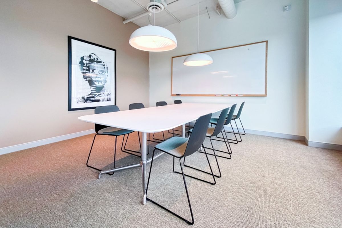 A modern office meeting room featuring a large whiteboard, pendant lighting, exposed ceiling ducting, and neutral carpeted flooring.