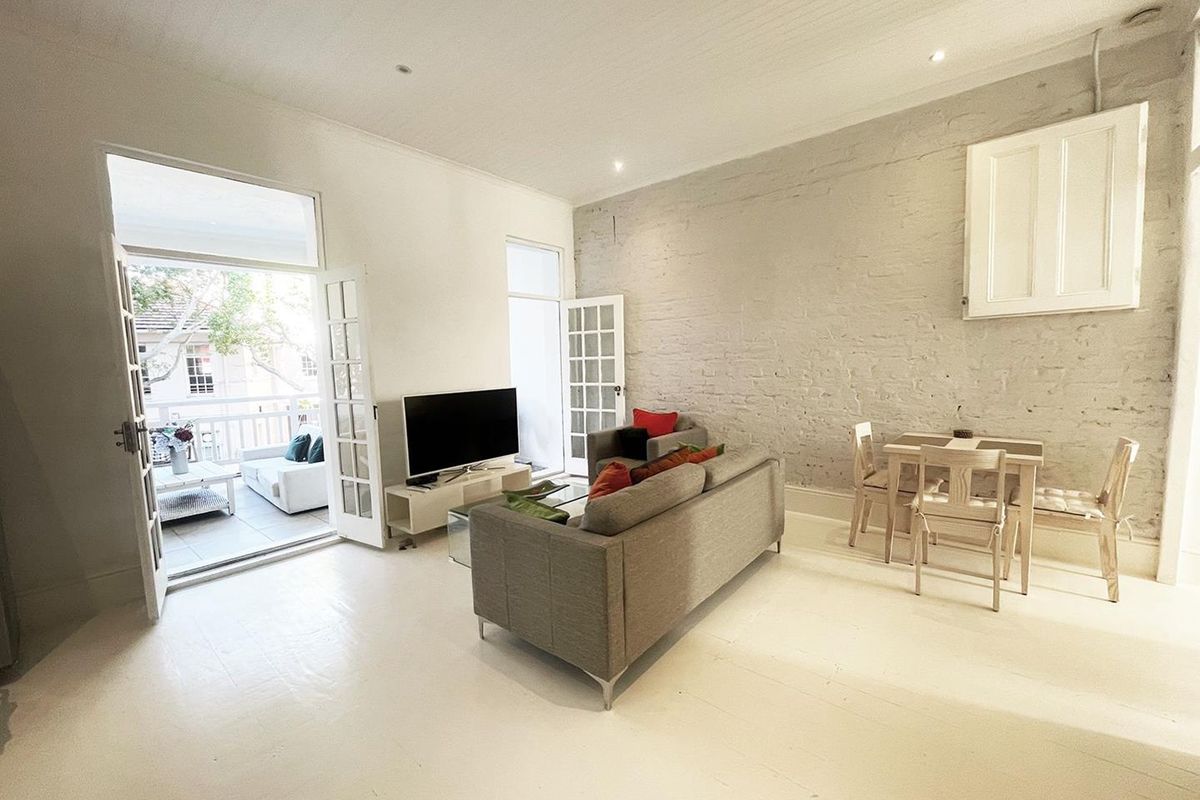 Bright living area featuring exposed brick walls, white timber flooring, and double french doors opening onto a private balcony.