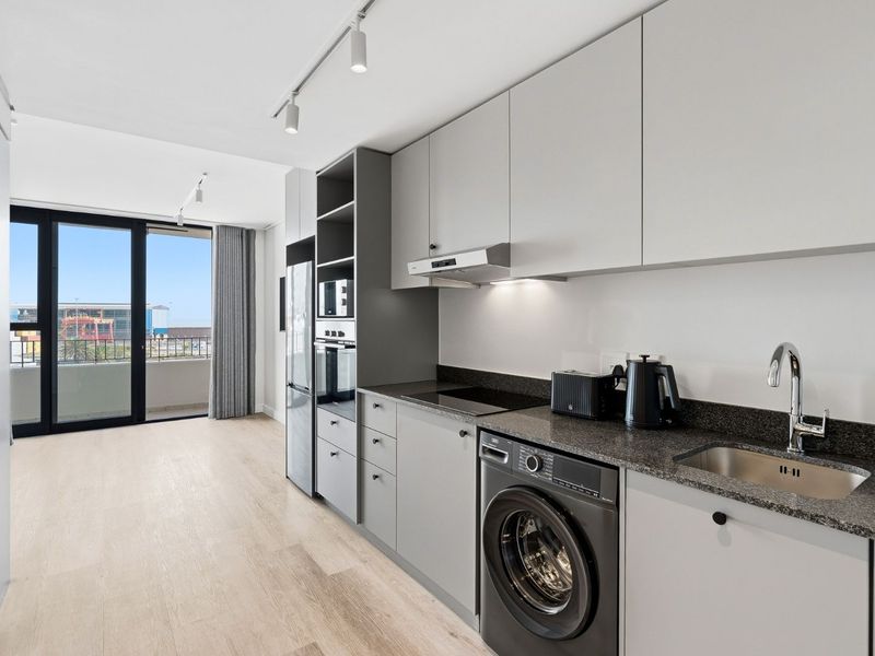 Modern open-plan kitchen featuring sleek grey cabinetry, stone countertops, integrated appliances, and direct access to a balcony through sliding glass doors.
