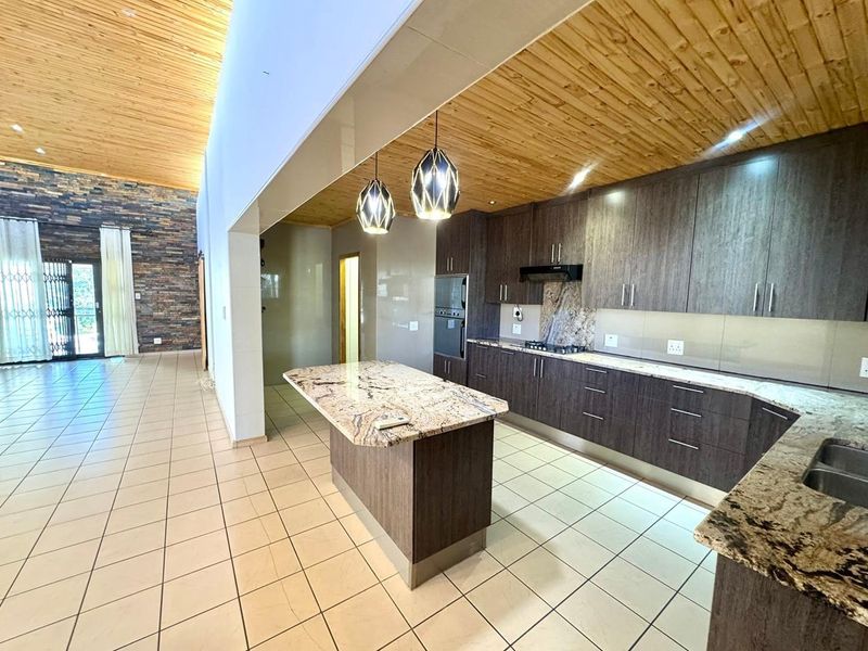 Spacious open-plan kitchen featuring dark cabinetry, granite countertops, a central island, and a striking timber-clad ceiling, seamlessly flowing into the living area.