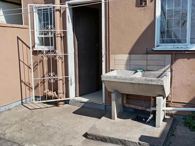 Rear exterior area featuring a concrete laundry trough, security gate, and exposed plumbing against a weathered building facade.