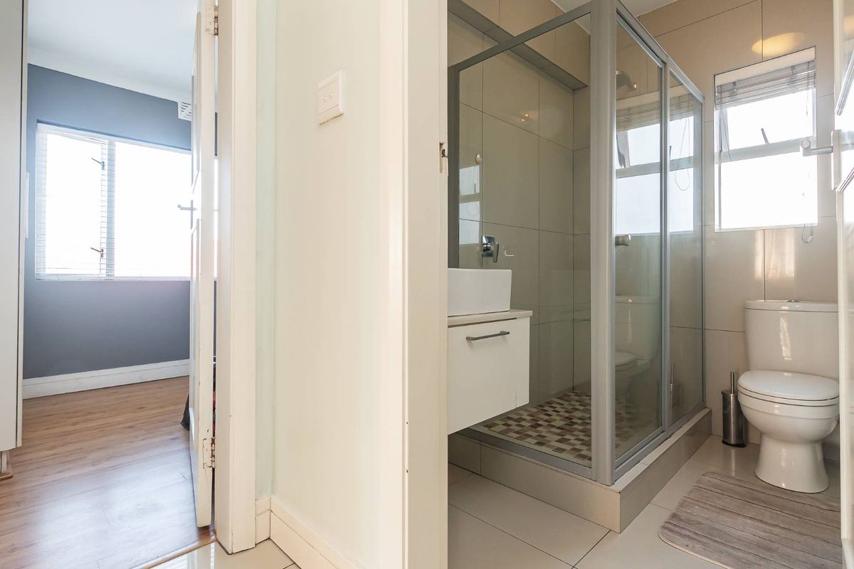 A modern bathroom featuring a glass-enclosed shower, vanity, and toilet, adjacent to a bedroom with wood-look flooring.