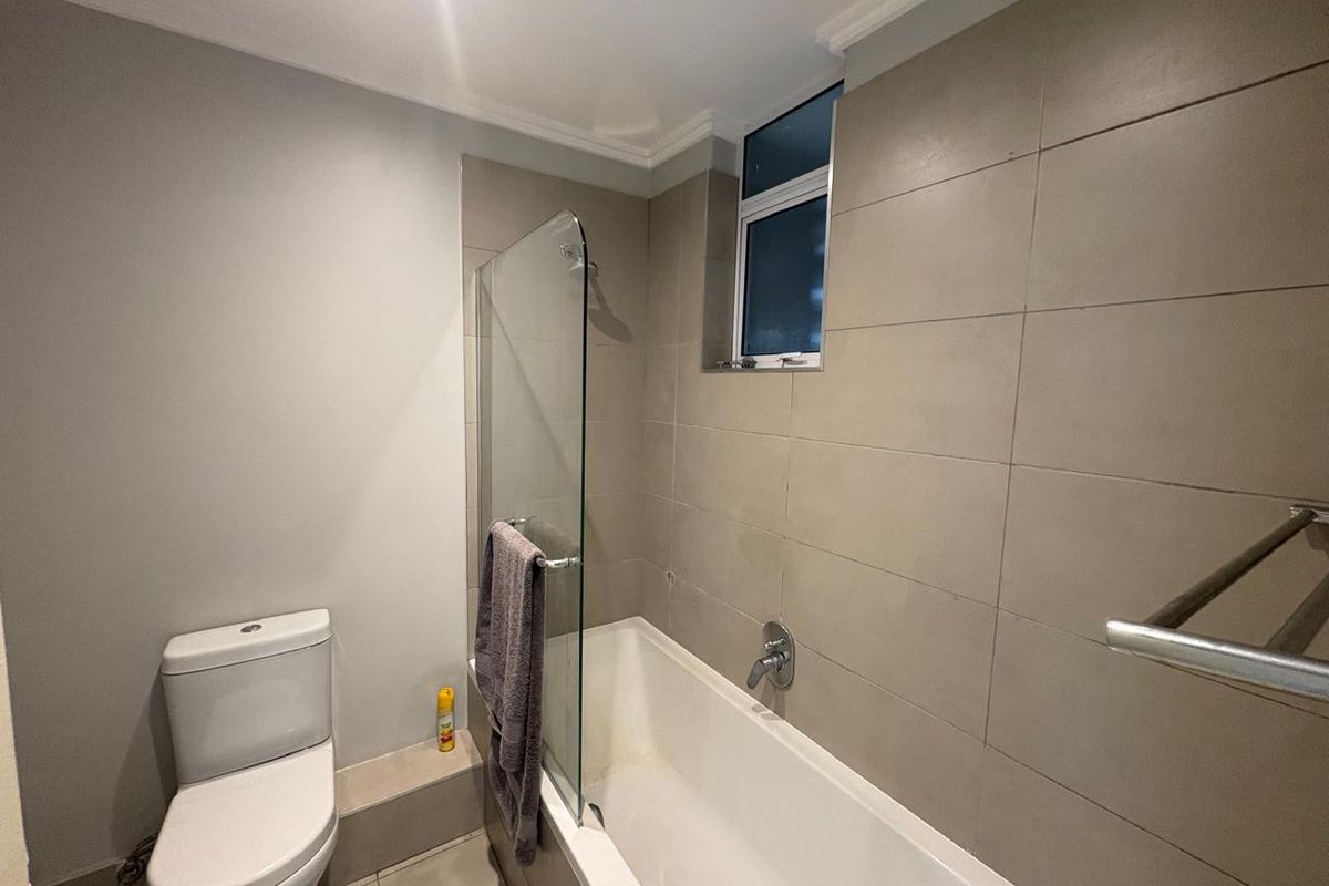 A functional bathroom featuring a bathtub with a glass shower screen, wall-mounted mixer, and a standard toilet with neutral wall tiling.