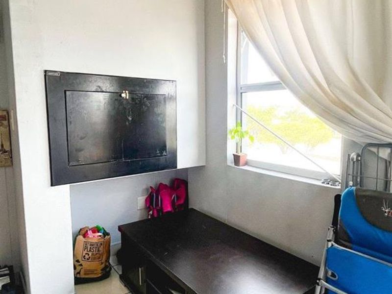 A small living area featuring tiled flooring and a window with curtains. The space includes a wall-mounted utility box and requires cosmetic updates.