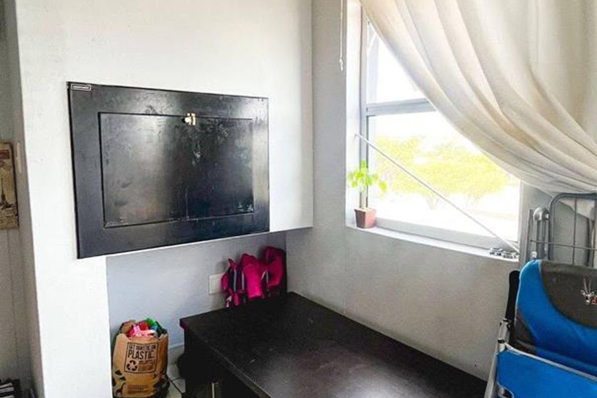 A small living area featuring tiled flooring and a window with curtains. The space includes a wall-mounted utility box and requires cosmetic updates.