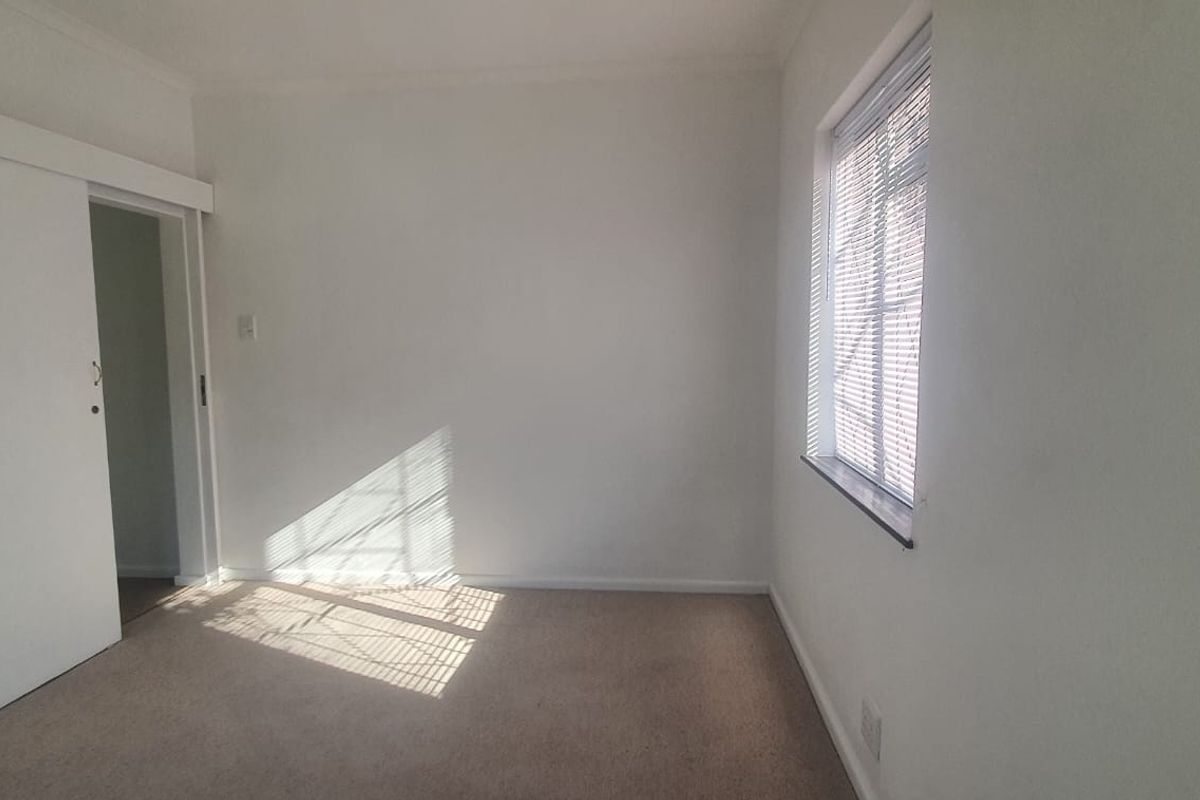 A bright, empty bedroom featuring neutral carpeted flooring, a sliding door, and a window fitted with security bars.