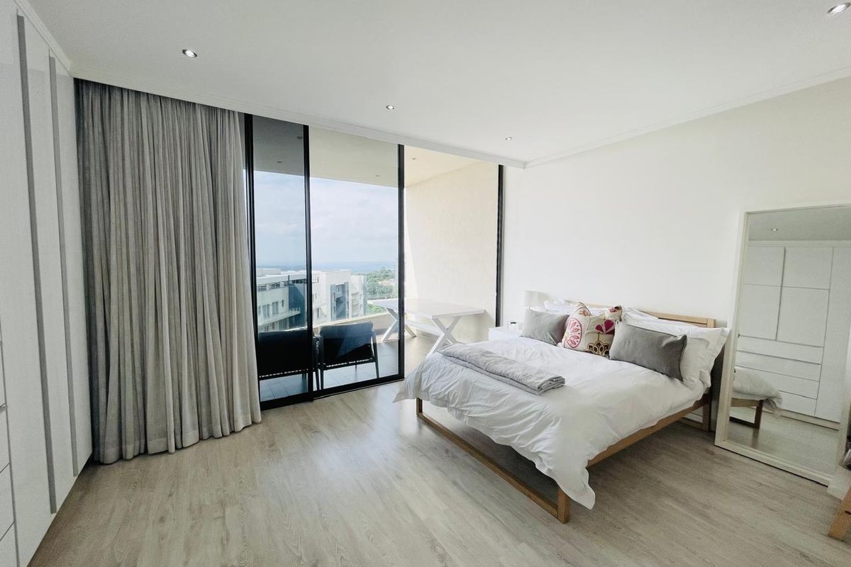 A bright, modern bedroom featuring light wood-look flooring, built-in wardrobes, and large sliding glass doors opening onto a private balcony with scenic views.