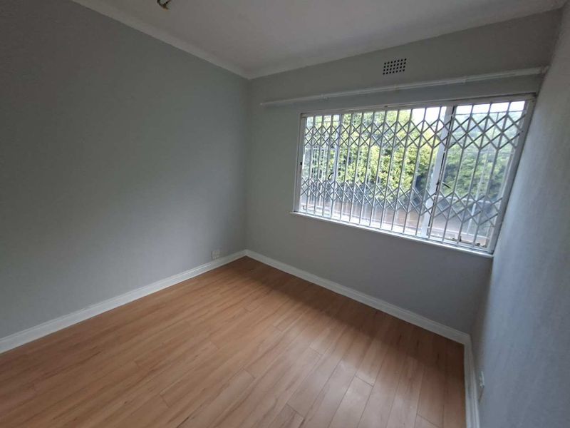 A bright, empty bedroom featuring light-toned laminate flooring, neutral walls, and a window fitted with a security gate.