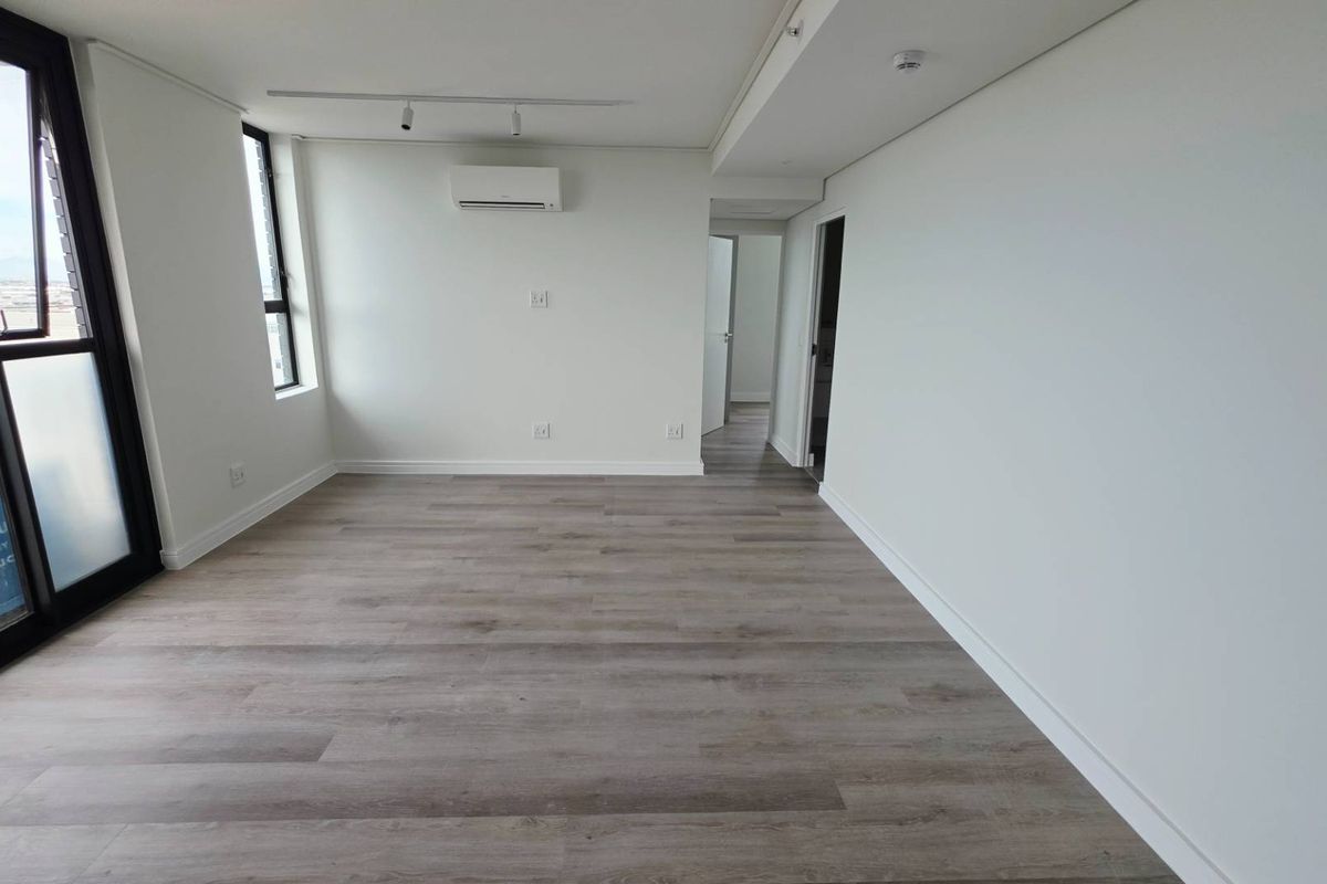 A bright, empty living room featuring modern laminate flooring, a wall-mounted air conditioning unit, and access to a balcony through a sliding glass door.