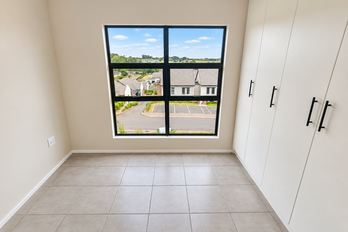 A bright, empty bedroom featuring neutral tiled flooring, modern built-in cupboards, and a large window offering a pleasant neighbourhood view.