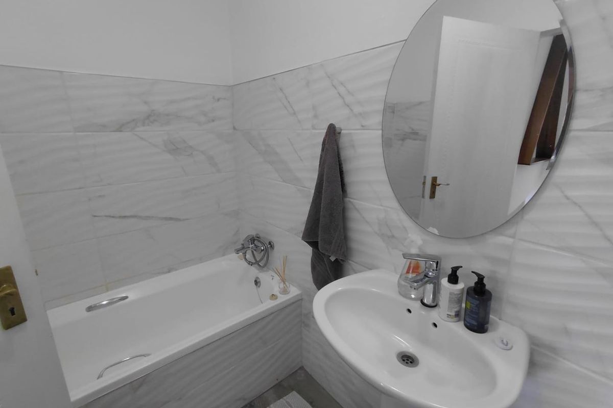 A clean, modern bathroom featuring marble-effect wall tiling, a white bathtub, and a pedestal basin with a large oval mirror.
