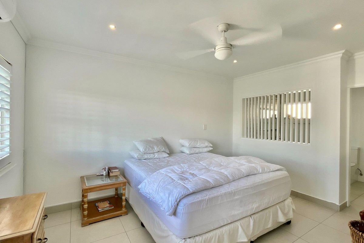 A bright bedroom featuring tiled flooring, a ceiling fan, split system air conditioning, and plantation shutters, with an adjoining ensuite bathroom visible.