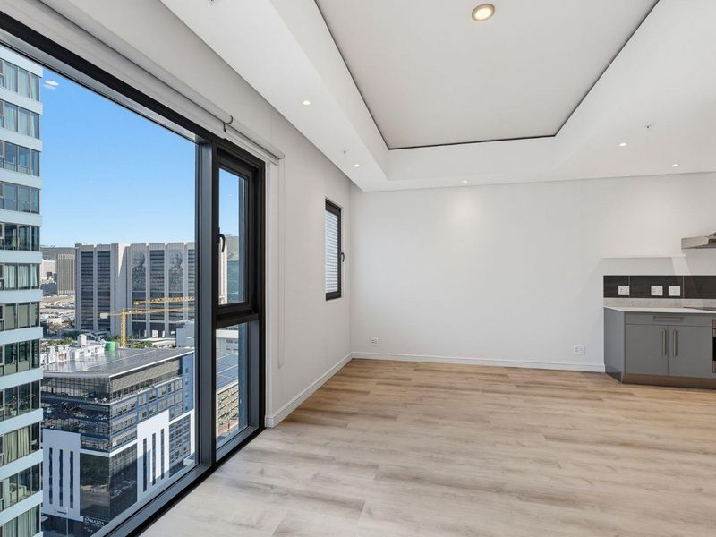 Modern open-plan living area featuring light laminate flooring, recessed ceiling lighting, and a compact kitchen with built-in appliances and expansive city views.